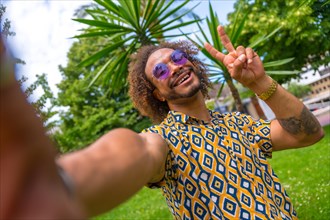 Afro-haired man on summer vacation next to some palm trees by the beach taking a selfie and doing the victory selfie. Travel and tourism concept