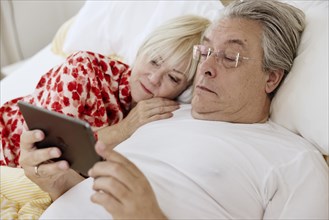 Elderly couple lying together in bed in bedroom looking at smartphone together