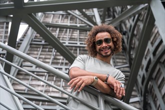 Portrait of a young man with afro hair wearing sunglasses on the stairs in the city smiling