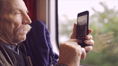 Elderly man traveling by train and taking pictures of the landscape through the window using a smartphone
