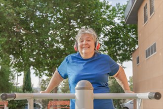 Active older woman in sportswear working out on a gym machine in the park while listening to music on her headphones and smiling at the camera