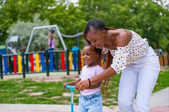 Black African ethnicity mother having fun with her daughter in playground learning to ride a skateboard