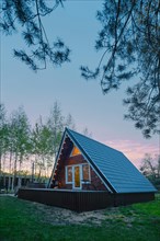 A-frame wooden cabin with light inside in early evening