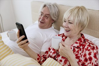 Elderly couple lying together in bed in bedroom looking at smartphone together