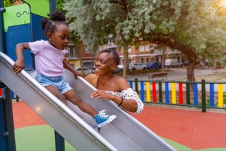 African black ethnicity mother having fun with her daughter in the squeaker of the playground of the city park in the sunset