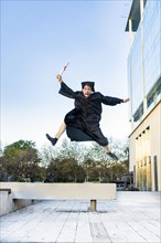 Happy graduated man in a black gown jumping while holding his diploma