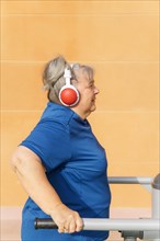 Profile view of an active obese elderly woman in sportswear working out on a gym machine in a park gym while listening to music on her headphones