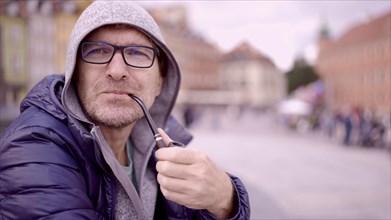 Portrait of adult man with glasses sitting in the hood on square and smoking a tobacco pipe in the Palace Square