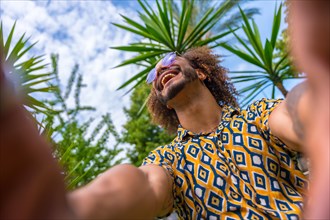 Afro-haired man on summer vacation next to some palm trees next to the beach taking a selfie with both hands smiling. Travel and tourism concept