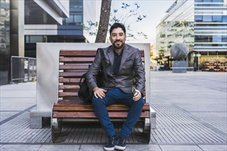 A businessman sitting on a bench in the street is looking at the camera with a smiling gesture