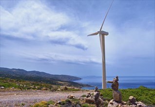 Single windmill turbine and balancing stones on hilltop of seashore in colorful landscape against dynamic blue sky with clouds and winding road on clear sunny summer day. Crete