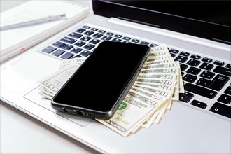 Cell phone on laptop keyboard and dollar bills. Mobile phone on top of dollar bills on laptop keyboard. Top view of laptop with cell phone and dollar bills