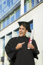 Happy graduated man wearing a bachelor gown and a black mortarboard and pointing at his diploma while looking at camera