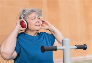 Active obese elderly woman in sportswear working out on a park machine while listening to music on her headphones