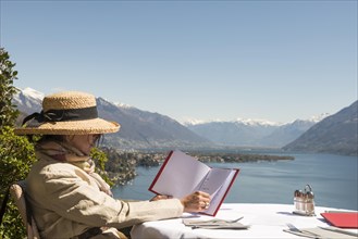 Woman with Straw Hat Sitting and Reading the Menu in a Restaurant with Panoramic View over Lake Maggiore in a Sunny Day in Ascona
