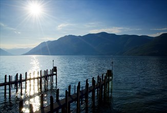 Pier with Sunbeam and Mountain on Alpine Lake Maggiore in Ticino