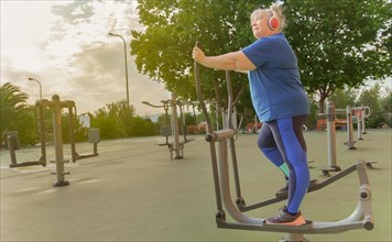 Active obese elderly woman in sportswear working out on a park machine while listening to music on her headphones