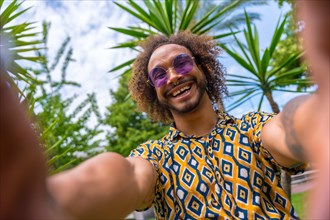 Afro-haired man on summer vacation next to some palm trees next to the beach taking a selfie with both hands smiling. Travel and tourism concept