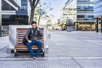 A businessman is talking on the cell phone while sitting on a bench in the street