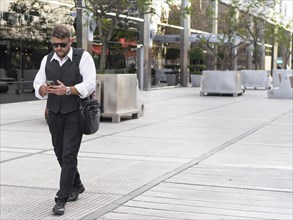 Portrait of a businessman walking on the street while he sends messages on his mobile phone