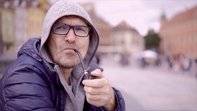 Portrait of adult man with glasses sitting in the hood on square and smoking a tobacco pipe in the Palace Square