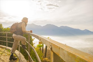 Woman with a Backpack Leaning on a Railing and Enjoy the Panoramic View over Locarno and Mountain in a Foggy Day in Ticino