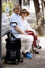 Older couple dressed in summer clothes with a trolley waiting for a bus at a bus stop