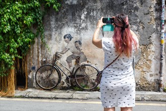 Young woman taking a photo of street art boy and girl on bicycle on a wall in George Town on Penang Island