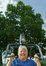 Older obese woman in sportswear exercising in an outdoor gym in a park surrounded by trees