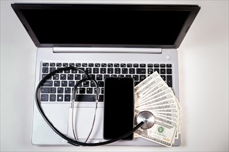 Mobile phone with dollar bills and stethoscope on laptop keyboard. Stethoscope on top of dollar bills on laptop keyboard