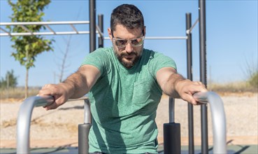 Man with beard and sunglasses training his arms on parallel bars in a park gymn