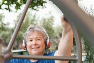 Active obese elderly woman in sportswear working out on a park exercise machine while listening to music on her headphones