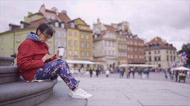 Elderly lady sits on the steps drinking coffee and using a smartphone in the historic center of an old European city. Palace Square