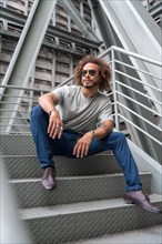 Young man with afro hair wearing sunglasses on the stairs in the city sitting looking to the left