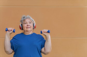 White haired obese older woman exercising with dumbbells and headphones listening to music for weight loss .isolated on orange background and copy space