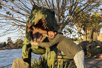 Latin man at an amusement park making himself enter the mouth of a fake dinosaur
