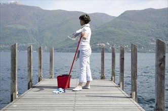 Woman Scrubs the Pier over the Alpine Lake with Cleaning Equipment and Mountain in Background in Ticino
