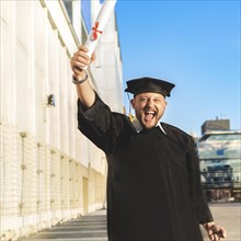 Happy graduated adult man wearing a bachelor gown and a black mortarboard and showing his diploma while looking at camera
