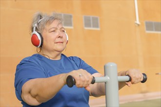 Active obese elderly woman in sportswear working out on a park machine while listening to music on her headphones