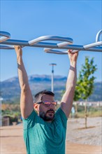 Bearded man with sunglasses seen from the front training his back by doing pull-ups on a barbell assisted by an elastic fitness band in an outdoor gym
