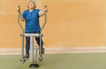 Active obese elderly woman in sportswear working out on a park machine while listening to music on her headphones