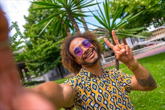Afro-haired man on summer vacation next to some palm trees by the beach taking a selfie and doing the victory selfie. Travel and tourism concept