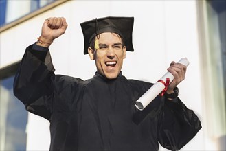 Happy young man wearing graduation robe holding diploma while raising his arm