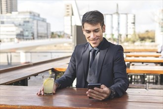 Businessman sitting at an outdoor bar drinking lemonade looking at his phone smiling