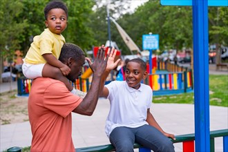 African black ethnicity father waving with his children in the playground of the city park