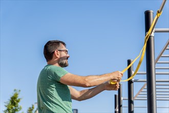 Bearded man with sunglasses seen in profile training with an elastic fitness band in an outdoor gym