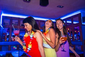 Female friends in a nightclub dancing with glasses of alcohol smiling at a night party on summer vacation