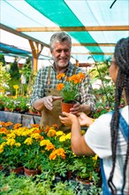 Happy female customer buying orange flowers from a gardener in a nursery inside the greenhouse