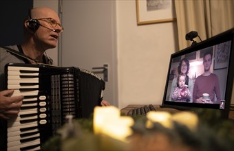 Christmas in times of great distance. Grandfather with accordion in front of a screen showing his family.