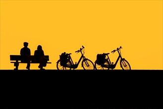 Couple of elderly cyclists resting on bench next to their two bicycles silhouetted against yellow sunset sky in summer